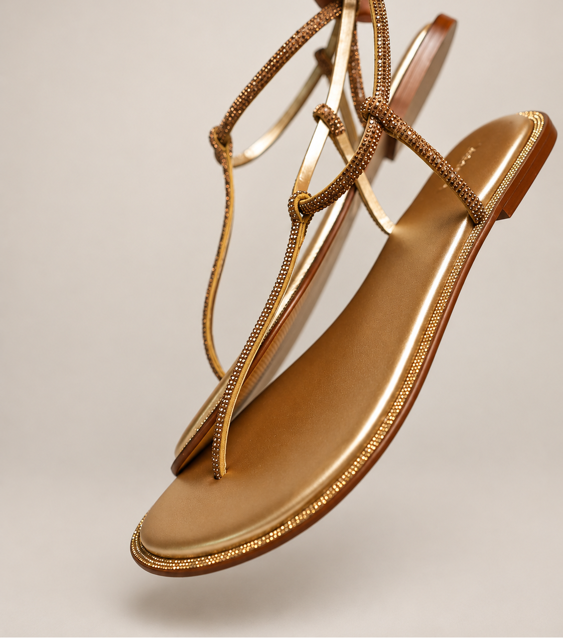A pair of metallic gold flat thong sandals with braided embellished straps, held by a hand against a neutral light gray background.