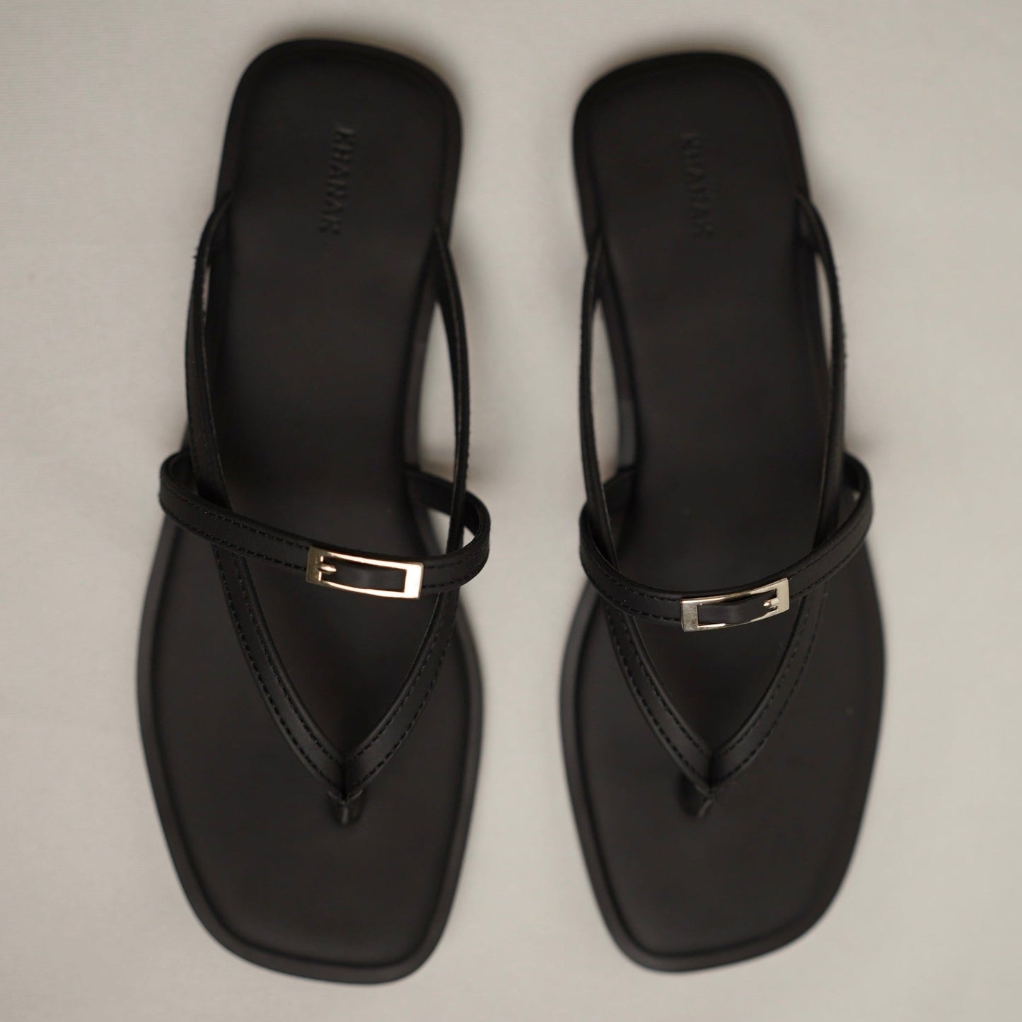 A pair of black thong sandals with square toes and a small silver metal accent on the strap, shown from a top-down view on a light gray background.