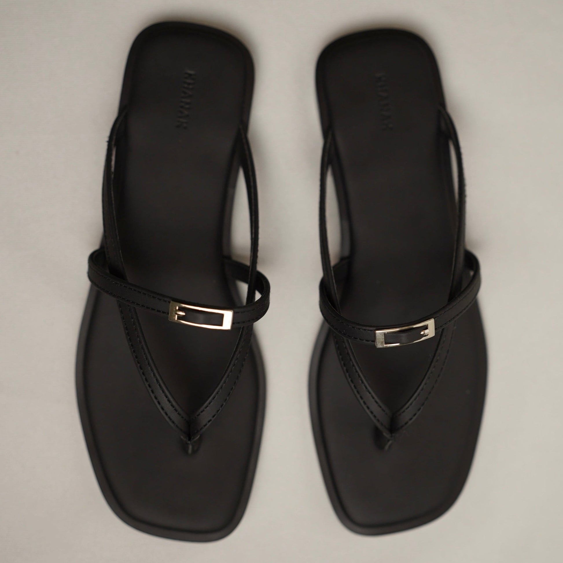 A pair of black thong sandals with square toes and a small silver metal accent on the strap, shown from a top-down view on a light gray background.