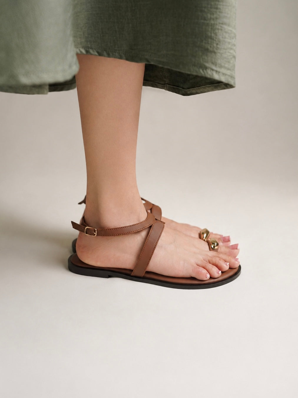 A person wearing a brown leather flat sandal with a toe loop and small gold-tone decorative accents, shown against a plain light beige studio background.