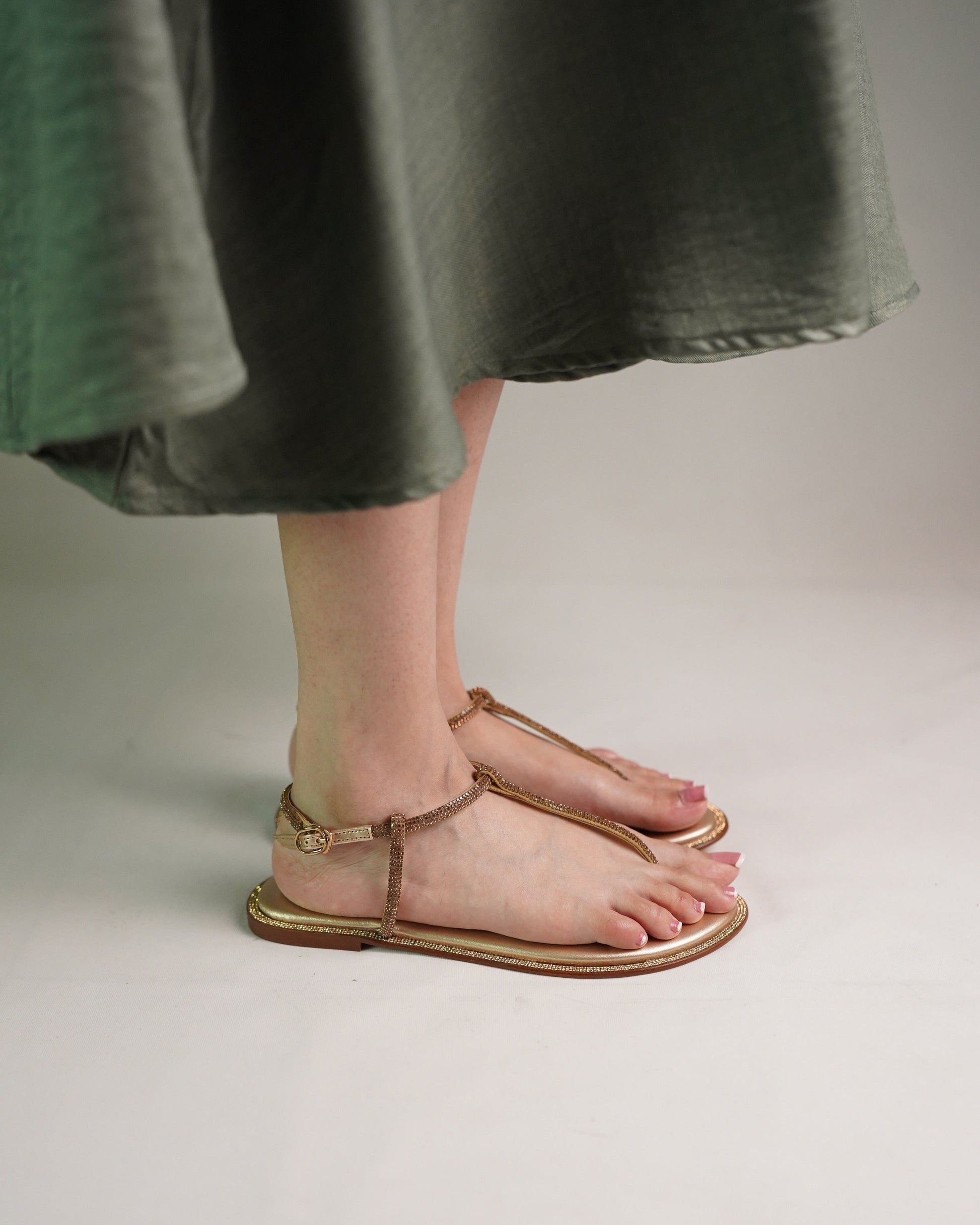 A person wearing a dark green skirt and bare feet in metallic gold flat sandals with ankle straps, standing against a light gray studio background.